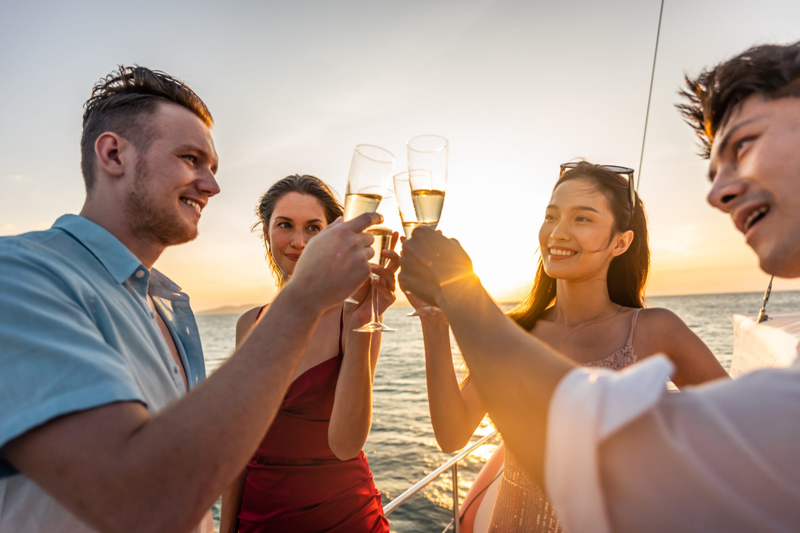Group of diverse friends drink champagne while having a party in yacht. Attractive young men and women hanging out, celebrating holiday vacation trip while catamaran boat sailing during summer sunset.